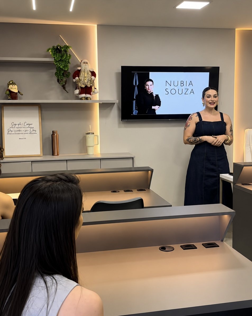 Núbia em pé, dando aula para as alunas na sala de curso, com o nome dela na TV