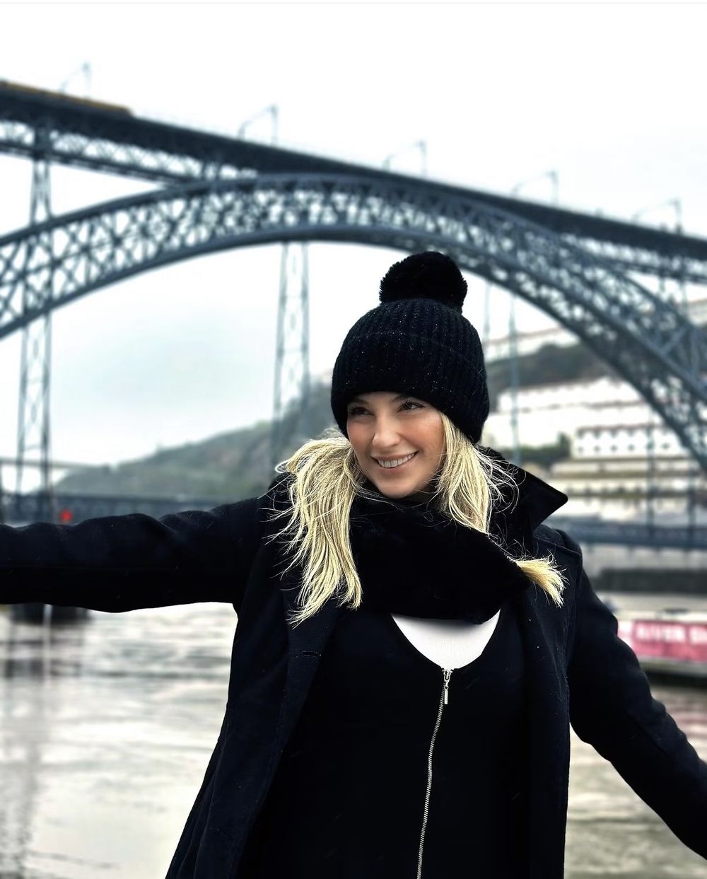 Núbia sorrindo de touca preta em frente à Ponte Dom Luís I, no Porto